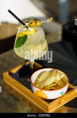 Erfrischendes Limoncello-Spritz mit Eis, Zitronenscheiben, Minze und Oliven im Glas, serviert mit knusprigen Kartoffelchips auf Holztablett, Sommergetränk-Konzept Stockfoto