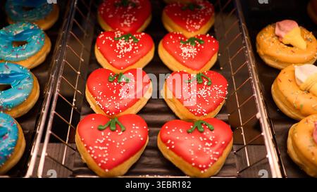 Herzförmige Donuts mit roter Glasur und Streuseln zum Valentinstag. Auswahl an bunten glasierten Backwaren auf einem Tablett in einer Bäckerei. Stockfoto