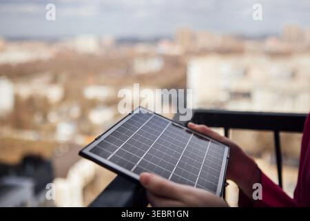 Die Person hält ein kompaktes Solarmodul auf einem Balkon, im Hintergrund ist eine Skyline der Stadt zu sehen, die nachhaltige Energielösungen im städtischen umfeld betont Stockfoto