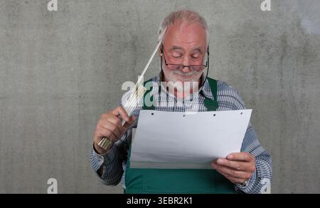 Verwirrter Senior Handwerker mit Blaupausen und faltbarem Lineal an einer Betonwand. Authentisches, humorvolles Porträt eines erfahrenen Profis. Stockfoto