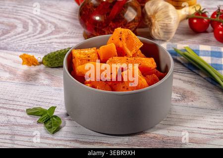 Gebackene, orangene Kürbisstücke, gewürzt mit Gewürzen in einer grauen Keramikschale. Gesundes vegetarisches Gericht auf hellem Holzhintergrund mit frischem Gemüse wie Bas Stockfoto