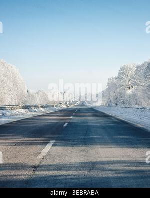 Lange, gerade Autobahn durch einen heiserfrostigen Wald. First-Person-Blick auf eine Straße, die von Bäumen umgeben ist, die mit weißem Raureif bedeckt sind. Stockfoto