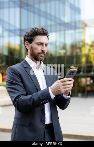 Reifer bärtiger Geschäftsmann mit dunklem Anzug und weißem Hemd, der draußen vor einem modernen Glasbürogebäude stand und sich auf sein Smartphon konzentrierte Stockfoto