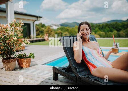 Junge Frau im Badeanzug, die am Pool sitzt und telefoniert. Stockfoto