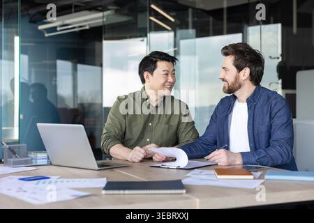 Zwei lächelnde multiethnische Geschäftskollegen prüfen Dokumente und arbeiten an einem modernen Bürotisch über einen Laptop zusammen, um Ideen und Planung auszutauschen Stockfoto