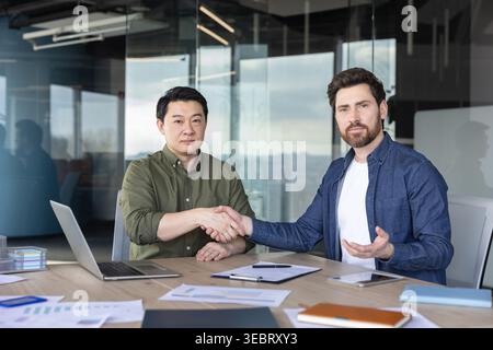 Verschiedene Geschäftspartner schütteln an einem hölzernen Konferenztisch in einem modernen Büro die Hand und symbolisieren erfolgreiche Partnerschaft, Zusammenarbeit, Stockfoto