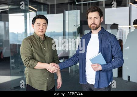 Verschiedene Geschäftsleute, ein bärtiger Mann und ein asiatischer Mann, der in einem modernen Büro die Hand schüttelt und für Partnerschaft, Vereinbarung und erfolgreiche Zusammenarbeit steht Stockfoto