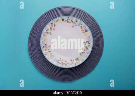 Overhead Shot verfügt über eine weiße Platte mit Blumenmustern, die auf einem grauen, gewebten Tischset aufliegt. Weiche, diffuse Beleuchtung hebt subtile Texturen und Vergangenheit hervor Stockfoto