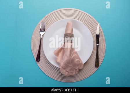 Top-Down-Kompositionsfunktionen Tabelleneinstellung. Teller mit pfirsichfarbener Serviette, Silberring, Gabel, Messer. Gewebte Matte. Blauer Hintergrund. Weiche, diffuse Beleuchtung Stockfoto