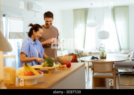 In einer hellen Küche kochen eine Koreanerin und ein Kaukasier zusammen. Sie bereitet Gemüse zu, während er lächelt und einen Drink hält. Sonnenlicht Stockfoto