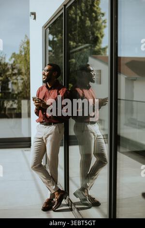Ein Mann steht an einem großen Fenster, schlürft an seinem Getränk, verlor in Gedanken, während er die friedliche Außenansicht bewundert, die in warmes Sonnenlicht getaucht ist. Stockfoto