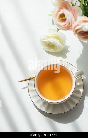 Elegante Lifestyle-Komposition mit weißer Keramik-Teetasse mit Muscheluntertasse und goldenem Rand, gefüllt mit bernsteinfarbenem grünem Tee, umgeben von blassrosa Stockfoto