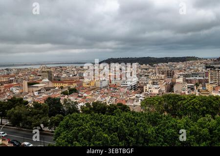 : Ein erhöhter Blick auf Cagliaris urbane Landschaft unter schweren grauen Wolken. Dichte Häuser, grüne Baumkronen und das ferne Meer bestimmen die Skyline. Stockfoto