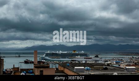 Ein großes Kreuzfahrtschiff und eine Fähre legen im Hafen von Cagliari an. Dunkle, stürmische Wolken hängen über dem Hafen und den fernen Bergen Sardiniens. Stockfoto