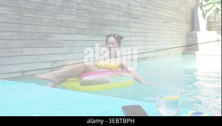 Liegende Frau in gelbem Badeanzug schwimmt auf einem bunten Ring am privaten Pool, Telefon Zitronenglas Stockfoto