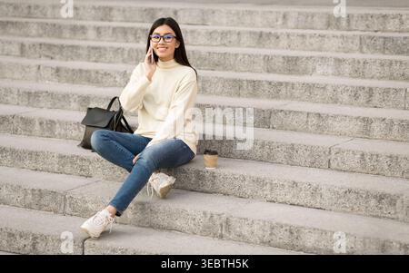 Junge Frau genießt einen Anruf, während sie mit Kaffee in der Hand auf Stufen sitzt Stockfoto