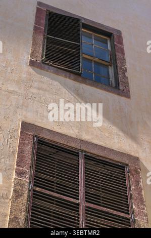 Zwei Fensterläden sitzen in einer sonnendurchfluteten Stuckwand auf Madeira. Obere Leisten sind halb über Glas geöffnet, untere Blenden geschlossen, gestreifte Schatten fallen im warmen af Stockfoto