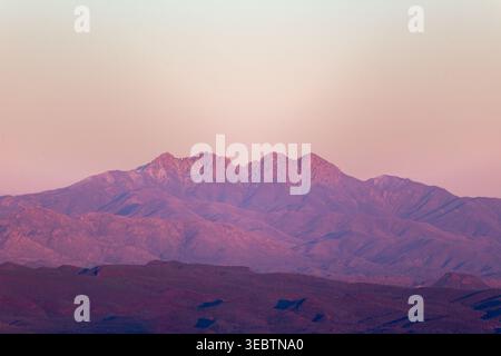 Weit entfernte Bergkette in sanftem rosa Abendlicht getaucht. Schroffe Gipfel stehen hoch vor einem trüben, warmen Himmel. Silhouetten von Graten erstrecken sich über den Horizont Stockfoto