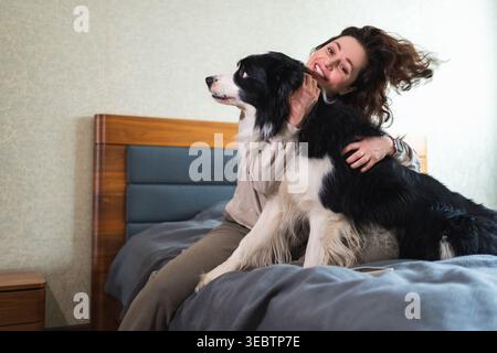 Lächelnde Frau, die mit niedlichem Hündchenhund Border Collie auf dem Bett zu Hause spielt. Besitzerin streichelt Hundefreund, der auf Sofa im Wohnzimmer sitzt. Liebes-Tier-Freundschaft-Team-Konzept Stockfoto