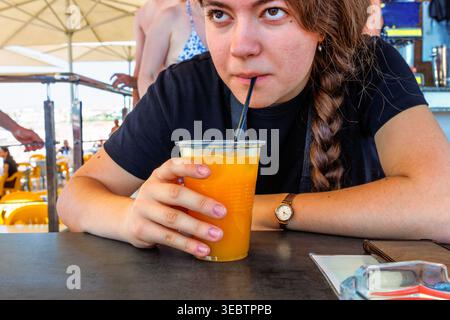 Junge Frau schlürft Orangensaft durch Strohhalm im Strandcafe mit Bikinigästen im Hintergrund. Paphos, Zypern am Mittelmeer, Stockfoto