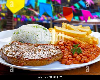 Brasilianisches Picanha-Steak mit weißem Reis, braunen Bohnen und knusprigen Pommes frites. Festlicher Hintergrund mit farbenfrohen Festa Junina Fahnen für Stockfoto