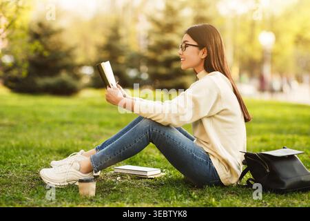 Die junge Frau liest gerne ein Buch über Gras in einem Sunny Park während des Frühlingnachmittags Stockfoto