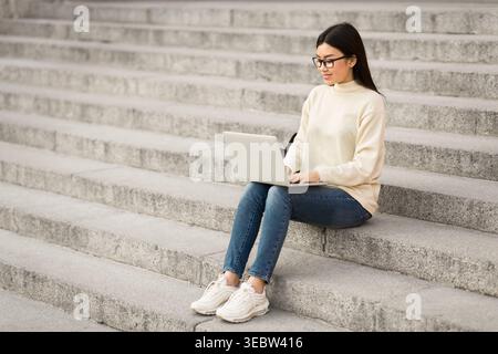 Die junge Frau arbeitet am Laptop, während sie in urbaner Umgebung auf Steintreppen sitzt Stockfoto