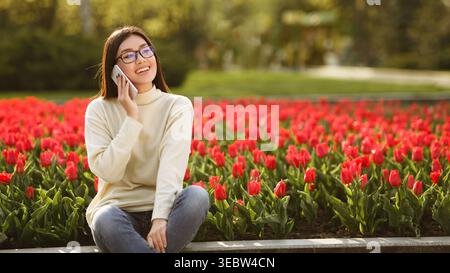 Junge Frau genießt einen sonnigen Tag, während sie zwischen blühenden Tulpen sitzt, während sie auf ihrem Telefon spricht Stockfoto