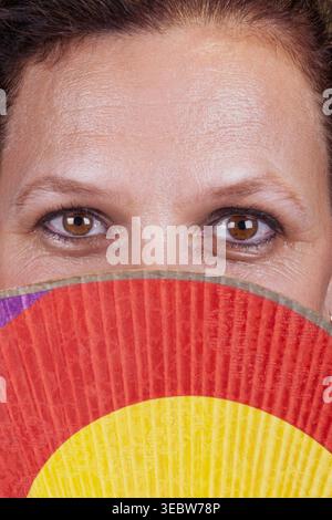 Nahaufnahme mit Blick auf die Augen einer Frau, die ihr Gesicht teilweise hinter einem leuchtend rot-gelben Faltenfächer versteckt Stockfoto
