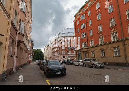 HELSINKI, FINNLAND - 11. AUGUST 2025: Von Bäumen gesäumte Stadtstraße mit geparkten Autos neben großen Wohnhäusern unter bewölktem Himmel. Stockfoto