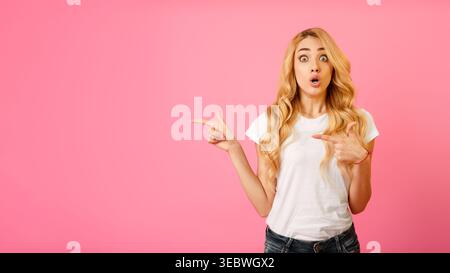 Junge Frau mit hellbraunem Haar posiert überraschend vor rosa Hintergrund, während sie zeigt Stockfoto