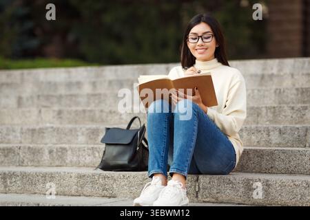 Junge Frau, die auf Stufen sitzt und in Notizbuch schreibt, während sie einen sonnigen Tag im Freien genießt Stockfoto