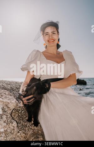 Frau Katze Küste fröhliche junge Dame in elegantem weißem Kleid und Hut mit einer schwarzen Katze an der felsigen Küste bei Sonnenuntergang Stockfoto