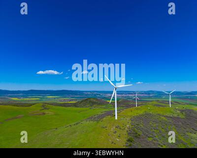 Windturbinen stehen hoch auf grünen Hügeln unter einem klaren blauen Himmel. Die Landschaft erstreckt sich in Richtung einer nahe gelegenen Stadt. Ein paar Wolken schweben über einem hellen Su Stockfoto