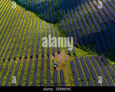 Eine Sammlung von Solarpaneelen erstreckt sich über Felder in einer Farm für erneuerbare Energien. Ein Traktor bewegt sich am helllichten Tag auf den Wegen zwischen den Blechen. Stockfoto