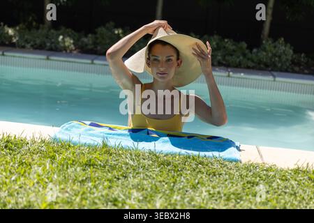 Eine mittelgroße Frau, die einen Sonnenhut anpasst, während sie im Pool im Hinterhof gelbe Badesachen mit blauem Handtuch trägt Stockfoto