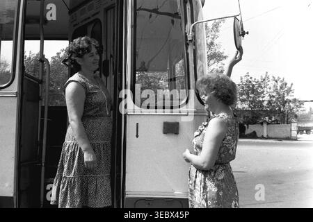 Slaviansk, 1986. Am Trolleybus-Depot. Zwei weibliche Mitarbeiter des öffentlichen Nahverkehrs interagieren in der Nähe der offenen Eingangstür eines Oberleitungsbusses. Eine Frau, die ein Pünktchenkleid trägt, steht auf der Treppe, während die andere in einem Blumenkleid den großen runden Seitenspiegel verstellt. Die Szene zeigt die tägliche Vorbereitung öffentlicher Fahrzeuge für die Stadtstrecke während der Sowjetzeit. Dieses authentische Archivfoto erfasst die Teamarbeit, die Wartung der Infrastruktur und die Arbeitsumgebung im historischen Kontext eines friedlichen Donbass. Stockfoto
