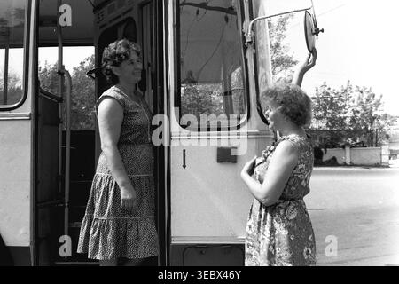 Slaviansk, 1986. Am Trolleybus-Depot. Zwei weibliche Mitarbeiter des öffentlichen Nahverkehrs interagieren in der Nähe der offenen Eingangstür eines Oberleitungsbusses. Eine Frau, die ein Pünktchenkleid trägt, steht auf der Treppe, während die andere in einem Blumenkleid den großen runden Seitenspiegel verstellt. Die Szene zeigt die tägliche Vorbereitung öffentlicher Fahrzeuge für die Stadtstrecke während der Sowjetzeit. Dieses authentische Archivfoto erfasst die Teamarbeit, die Wartung der Infrastruktur und die Arbeitsumgebung im historischen Kontext eines friedlichen Donbass. Stockfoto