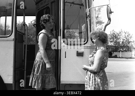 Slaviansk, 1986. Am Trolleybus-Depot. Zwei weibliche Mitarbeiter des öffentlichen Nahverkehrs interagieren in der Nähe der offenen Eingangstür eines Oberleitungsbusses. Eine Frau, die ein Pünktchenkleid trägt, steht auf der Treppe, während die andere in einem Blumenkleid den großen runden Seitenspiegel verstellt. Die Szene zeigt die tägliche Vorbereitung öffentlicher Fahrzeuge für die Stadtstrecke während der Sowjetzeit. Dieses authentische Archivfoto erfasst die Teamarbeit, die Wartung der Infrastruktur und die Arbeitsumgebung im historischen Kontext eines friedlichen Donbass. Stockfoto