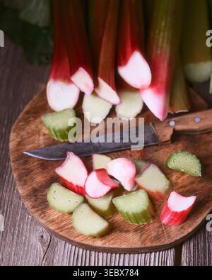 Gehackte Rhabarberstücke auf rundem Holzschneidebrett mit Messer, rustikaler Hintergrund. Frische, rohe Rhabarberscheiben in Nahaufnahme, Bio-Kochzubereitung Stockfoto