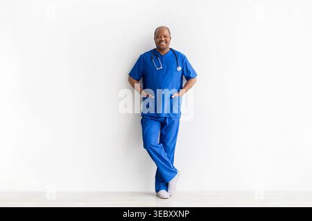 Selbstbewusster, reifer, männlicher Arzt in blauen Peelings, der sich an die weiße Wand lehnt Stockfoto