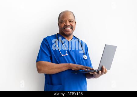 Glücklicher reifer afroamerikanischer männlicher Arzt in blauen Schutten, der Laptop hält Stockfoto