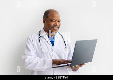 Glücklicher reifer afroamerikanischer männlicher Arzt im weißen Labormantel mit Laptop Stockfoto