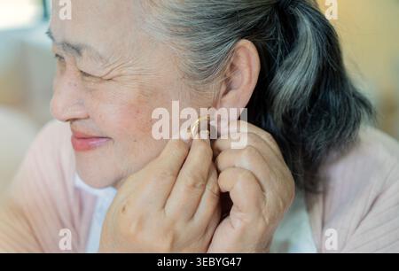 Eine ältere Frau, die einen Ohrring aufsetzt. Nahaufnahme einer glücklichen Seniorin, die den Ohrring aufsetzt. Die alte Dame probiert goldene Ohrringe an. Senio Stockfoto
