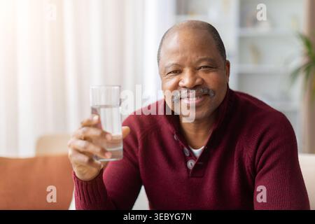 Lächelnder reifer schwarzer Mann, der ein Glas Wasser zu Hause hält Stockfoto