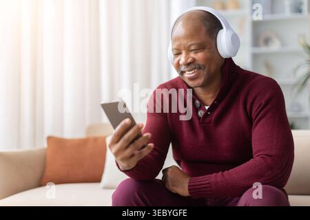 Reifer schwarzer Mann mit schnurlosen Kopfhörern und lacht, während er zu Hause das Smartphone benutzt Stockfoto