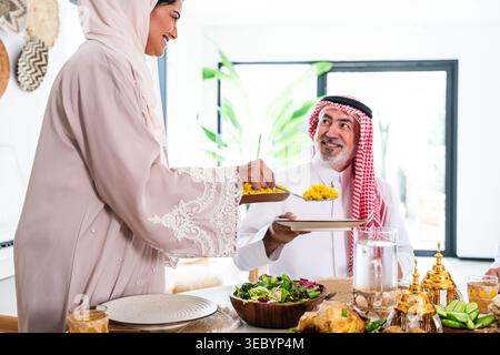 Arabische Frau, die einen Hijab trägt und einem lächelnden Mann traditionellen gelben Reis aus dem Nahen Osten serviert, in Kandura und Gutra an einem Familienesstisch mit Salat A Stockfoto