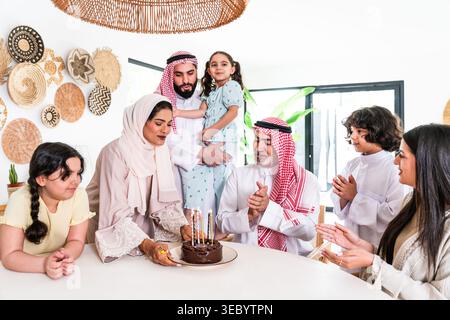 Die arabische Familie versammelt sich an einem Tisch, klatscht und lächelt, während eine Frau Kerzen auf einer Schokoladentorte anzündet, was eine fröhliche Atmosphäre für schafft Stockfoto