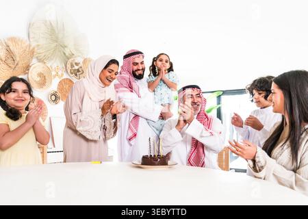 Arabische Großfamilie feiert zu Hause Geburtstag, fröhliches generationenübergreifendes Treffen um einen Schokoladenkuchen mit Kerzen, Klatschen, Lächeln und Stockfoto