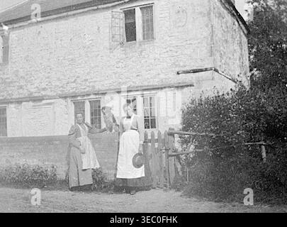 Ein einfarbiges Foto von Old Farm aus dem Jahr 1898 in Trowbridge, Wiltshire. Das Bild zeigt zwei Frauen mit Schürzen, die draußen neben einem Steingebäude stehen; eine hält ein kleines Tier auf einer Steinmauer, die andere steht in der Nähe eines hölzernen Tors. Aus einem späten viktorianischen Album mit kleinen Fotos, die von einem Amateurfotografen aufgenommen wurden. Die meisten stammen zwischen 1898 und 1901 und scheinen auf die Familie von John White (möglicherweise aus Sth London), einem Bauunternehmer und Bauunternehmer, zu beziehen. Die Qualität der Originalfotos war variabel und viele waren verblasst und verfärbt. Stockfoto